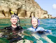 Two smiling snorkelers at the surface beside layered coastal cliffs